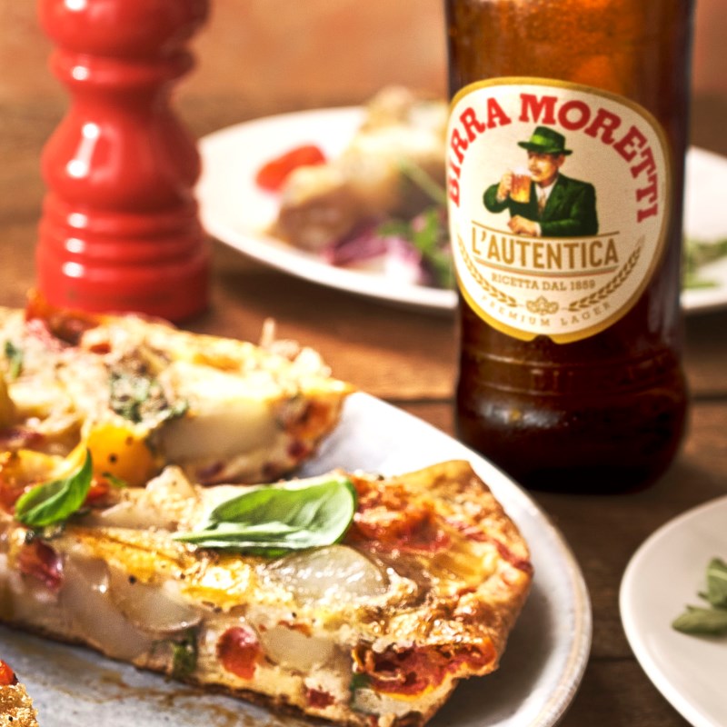 A slice of frittata garnished with basil leaves on a plate next to a Birra Moretti.